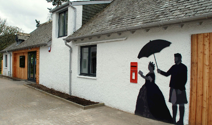 White building with slate roof with a black silhouette artwork on the side, depicting a woman in large dress and man holding an umbrella above