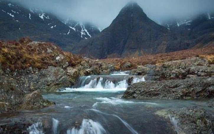 Small stream flowing towards camera through bog and heather covered ground with dark grey mountains beyond shrouded in cloud