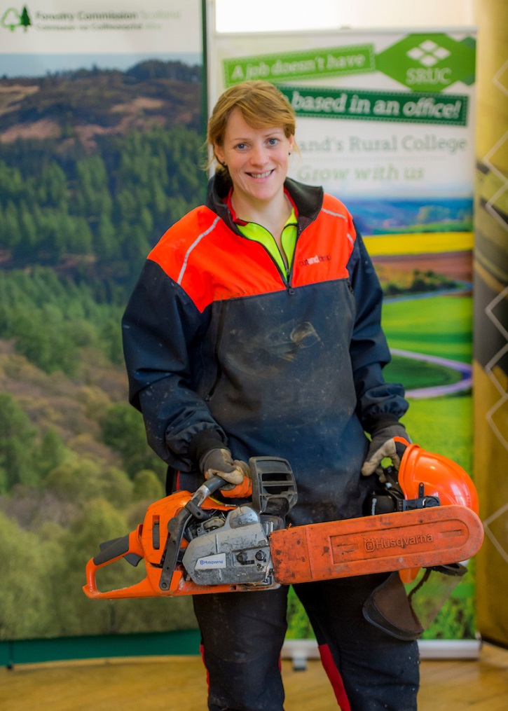 A woman in outdoor working clothes holding a chainsaw and smiling for the camera