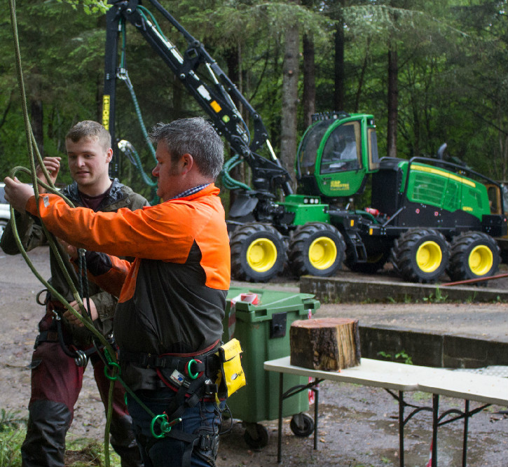 Man in harness demonstrating rope work to another man, with a forest forwarder parked in the background