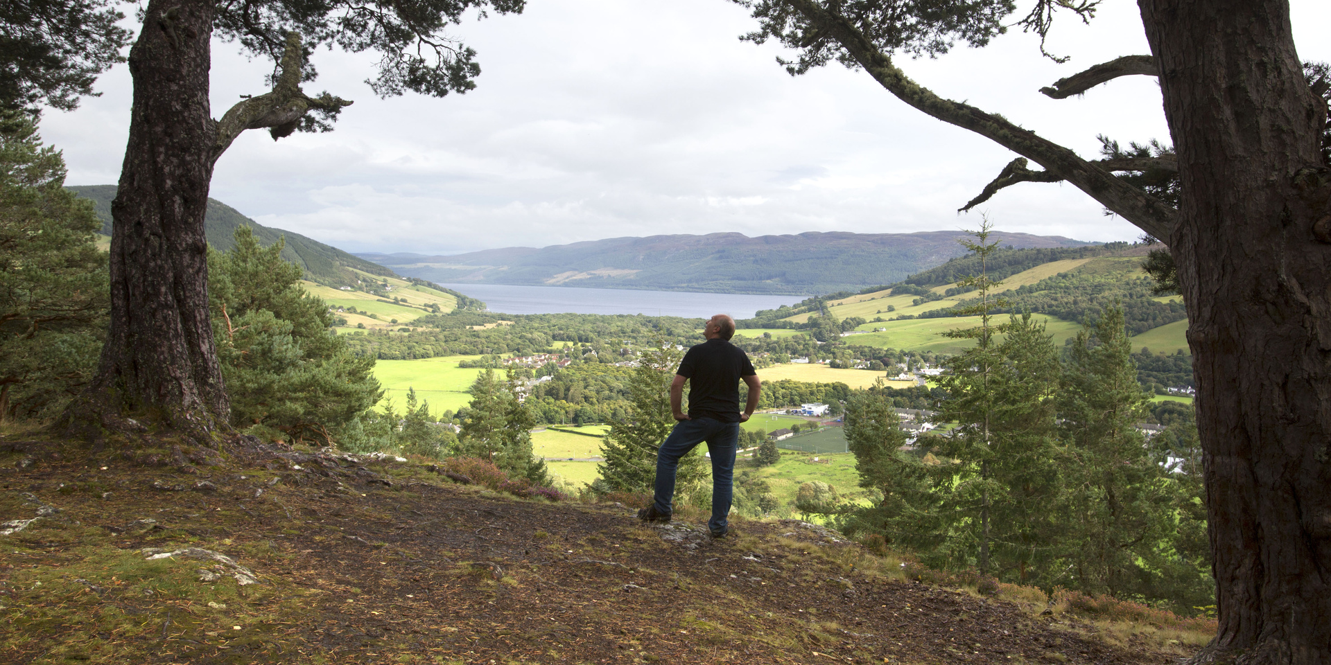 Man standing at hilltop viewpoint with trees, fields, loch and houses in the distance