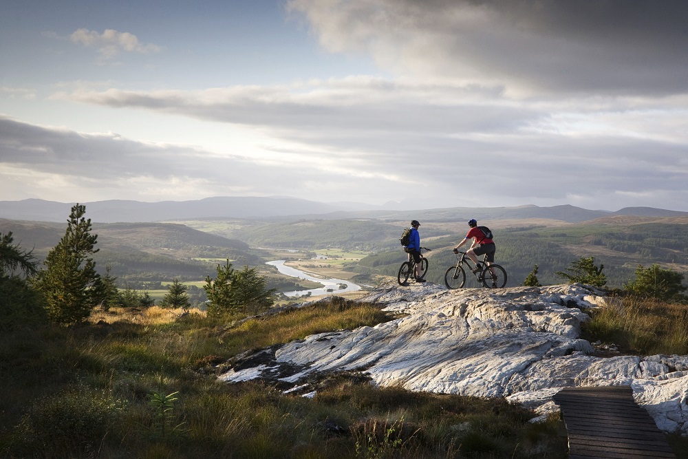 Two mountain bikers stopped at the top of a hill to look at a panorama of loch and hills