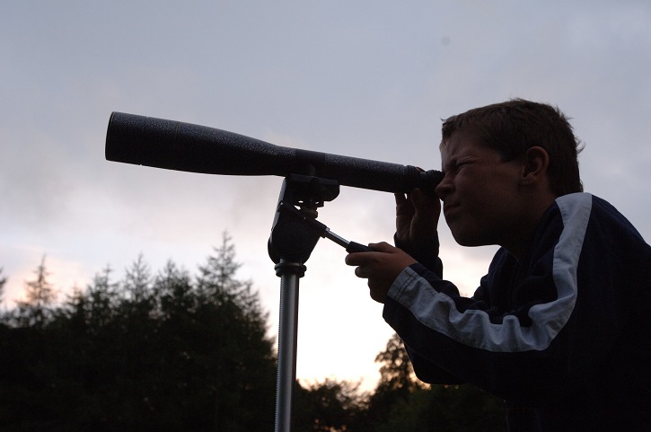 Boy with eye to stationary telescope with sky and trees beyond