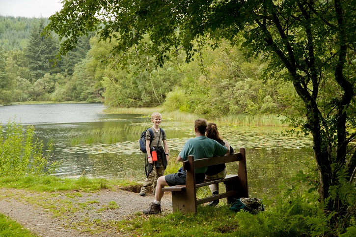 Two adults sitting on a bench with a child overlooking a loch surrounded by green-leaved trees