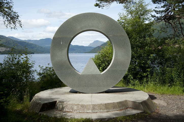 Large round sculpture or monument in front of a viewpoint over a loch surrounded by trees