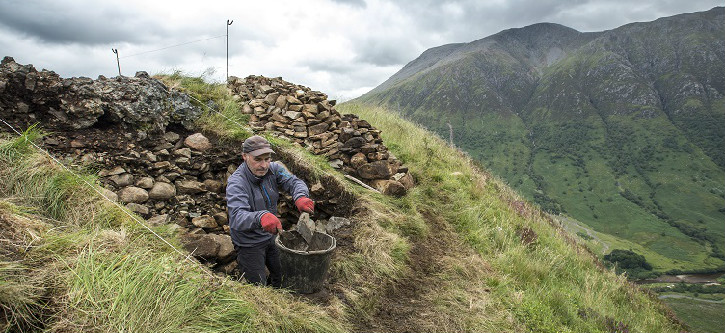 Man working to build stones into a wall high on a grassy hill with bigger hill in background