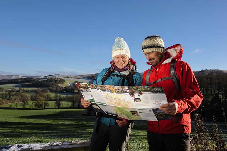 Two people in jackets, hats and rucksacks inspecting a map under a blue sky with green countryside beyond