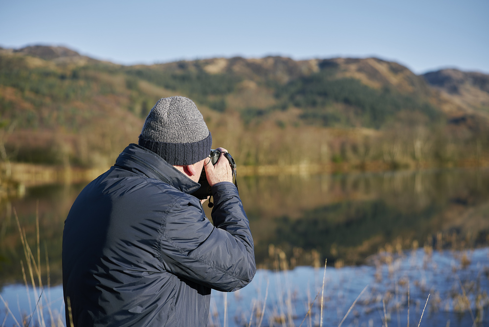 Man in jacket taking a photo of a calm loch with camera