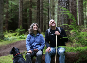 A man, a woman and a dog sitting on a fallen tree in a forest