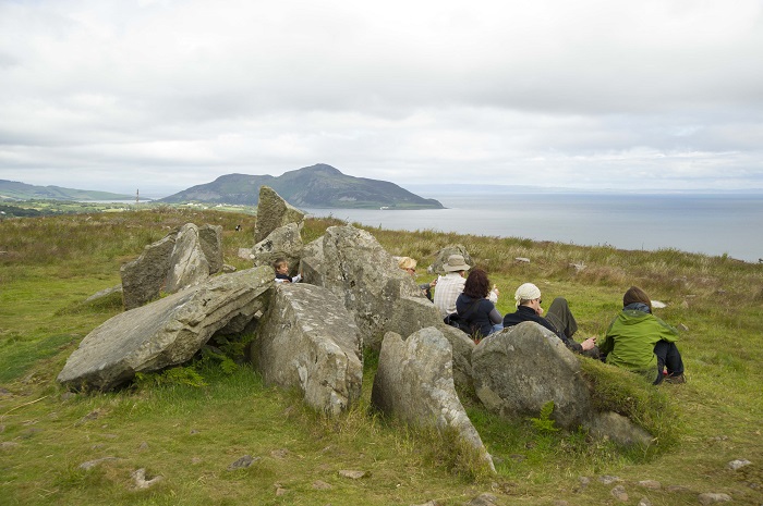 Several people sitting on the grass surrounding several large, fallen stones with a view out to sea
