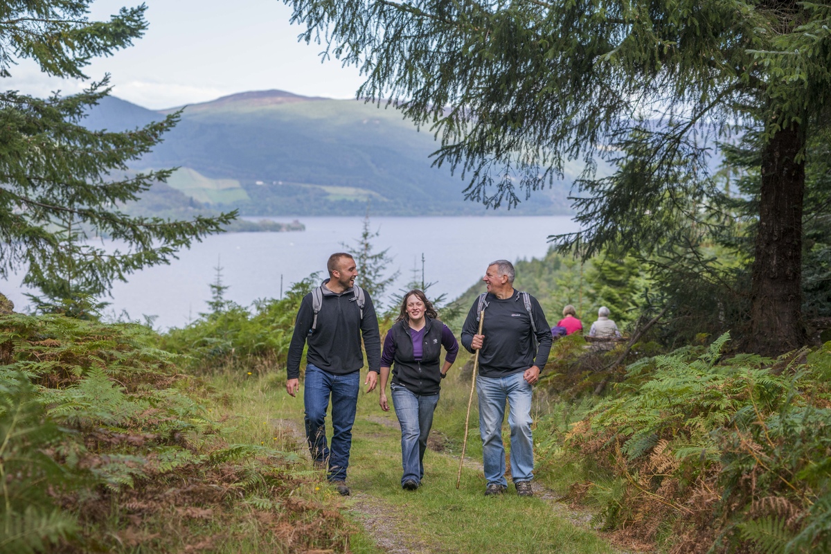 Two men and a woman walking along a woodland path with a large loch in the background