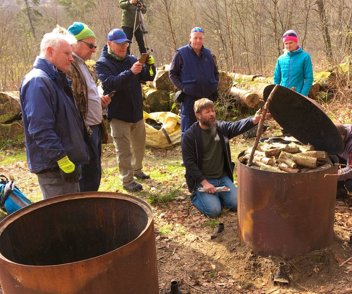 Several adults gathered around a large metal barrel packed with wood