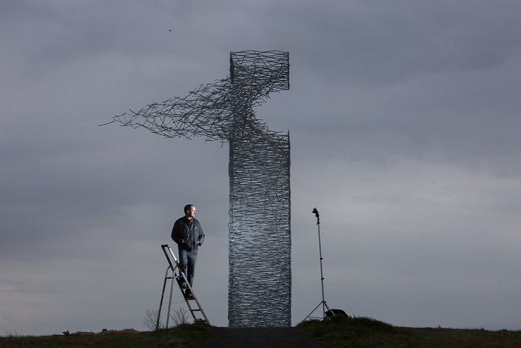 Man standing on step ladder outdoors in front of large see-through sculpture with grey cloudy sky behind