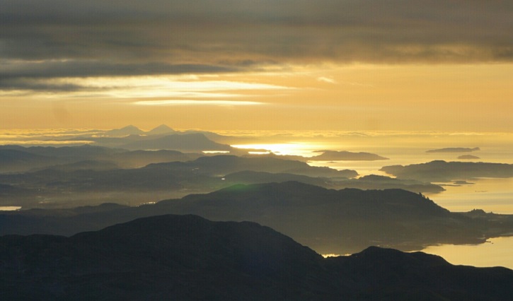 View from a mountain looking out over the landscape to sea during sunset, showing several silhouettes of other hills