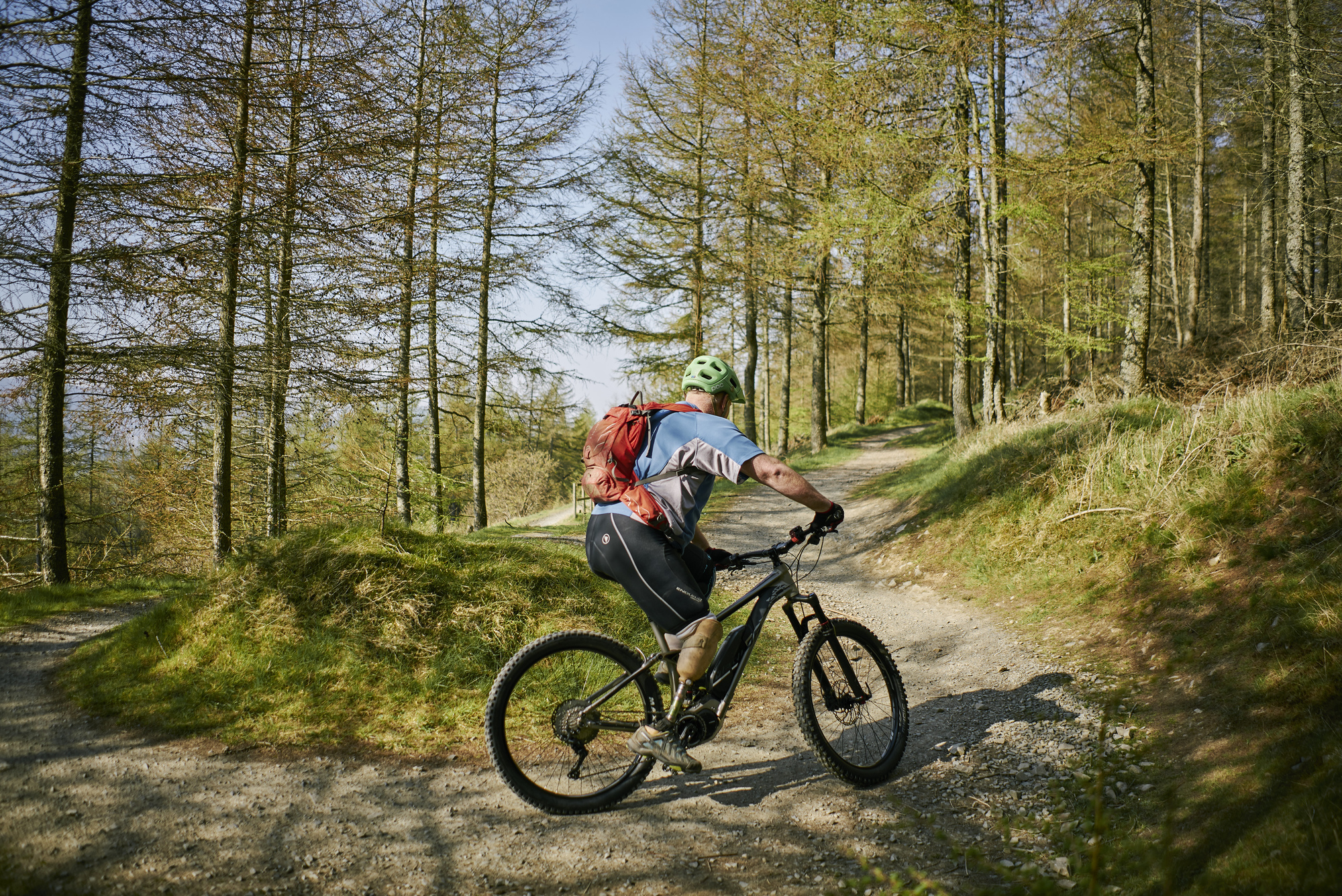 Male mountain biker (with prosthetic leg), dressed in blue T shirt, green helmet and orange rucksack, cycles round bend up hill on elevated stony track in Glentress Forest, near Peebles