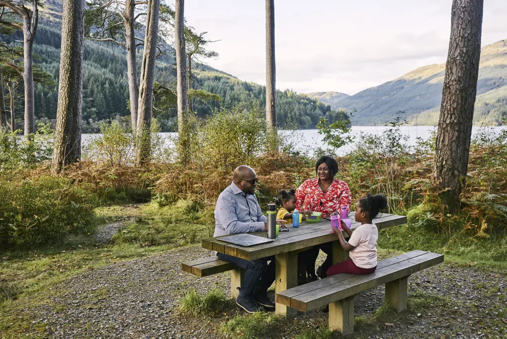  Man, woman and two young girls sit at picnic table with food and drink, at Jubilee Point, on shore of Loch Eck