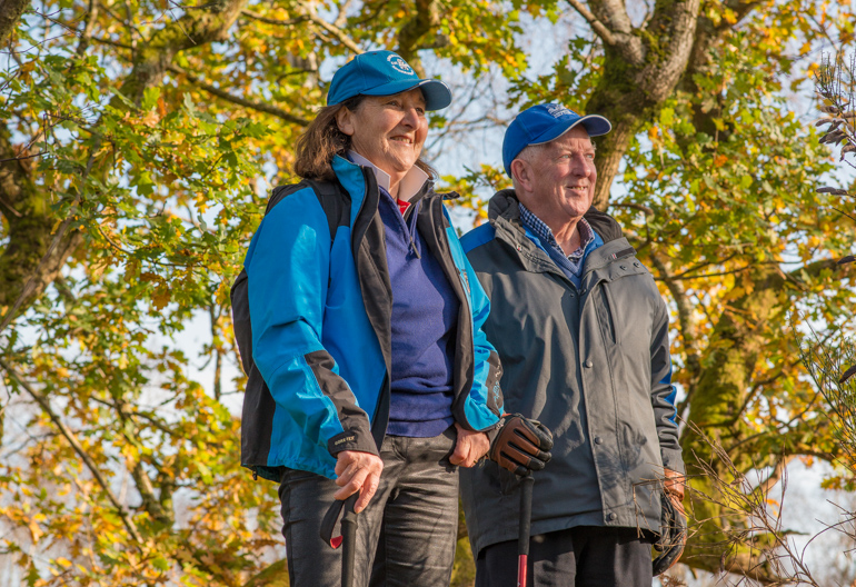 Man and woman in jackets and caps standing in front of some trees admiring a view.