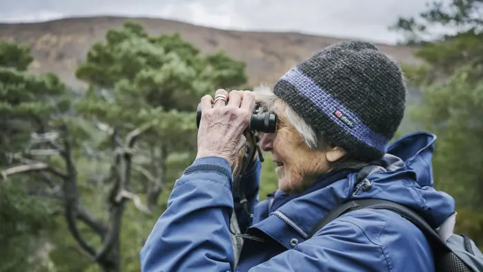 Person with binoculars with pine tree and hill in the background.