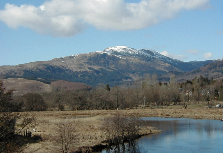 A small loch in winter sitting at the foot of a mountain with a bare forest and clouds in the reflection of the water