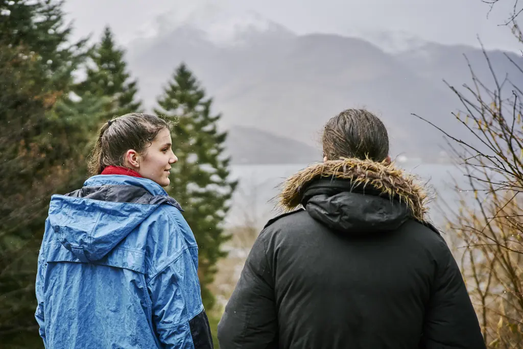 Young woman wearing blue waterproof jacket and young man in black jacket, stand, on a rainy day, at viewpoint near Tom a Chomrain looking over Loch to misty snow capped Pap of Glencoe mountain.