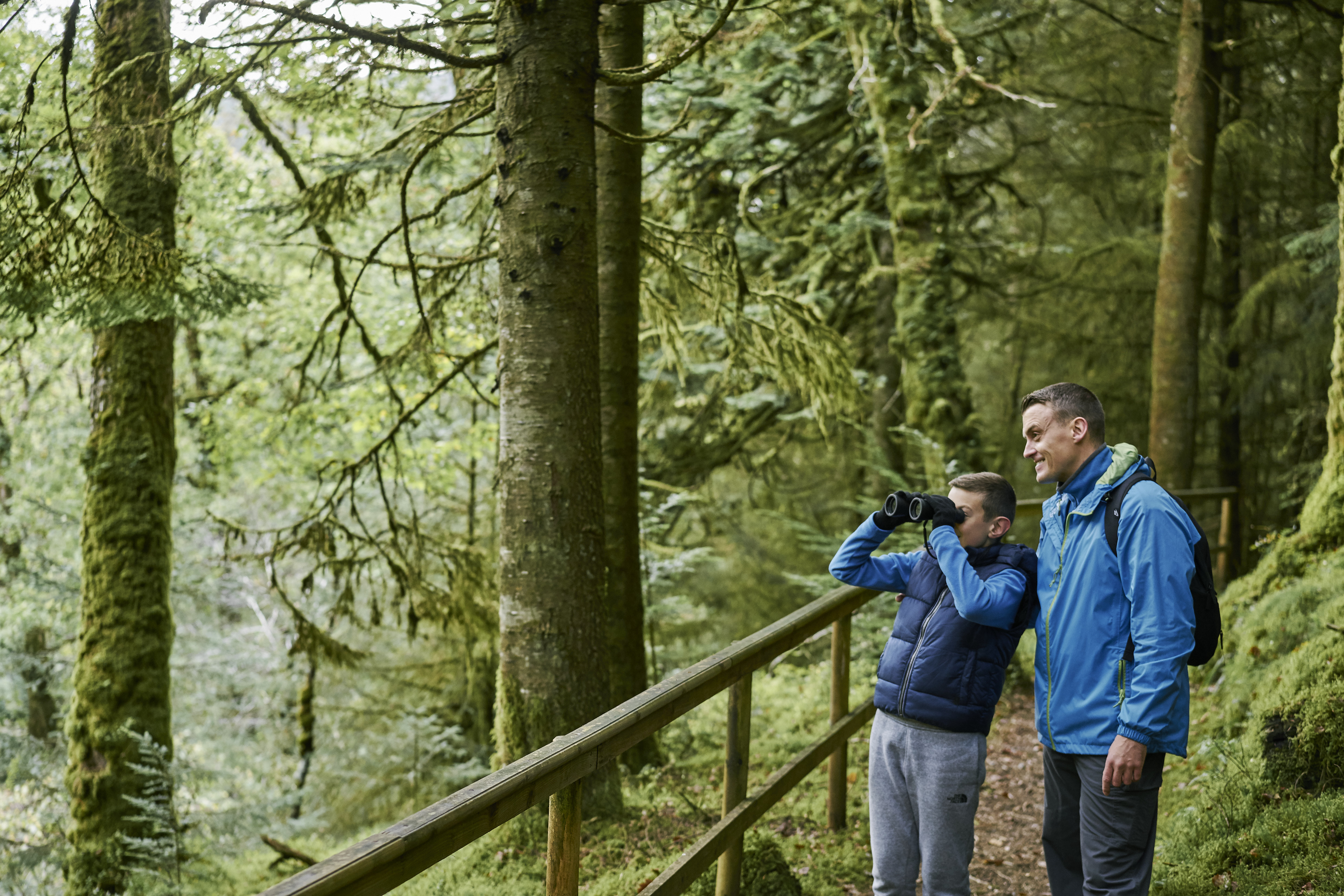a boy with binoculars and a man stand in a forest looking to the left