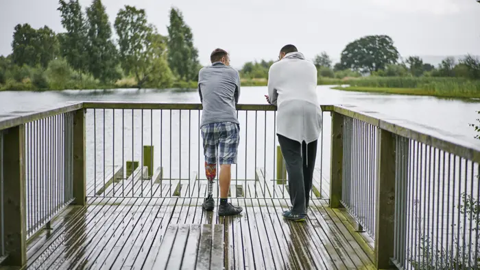Rear view of two men (one with prosthetic leg) standing on wooden pier looking across pond, in Larbert Woods in grounds of Forth Valley Royal Hospital, Larbert