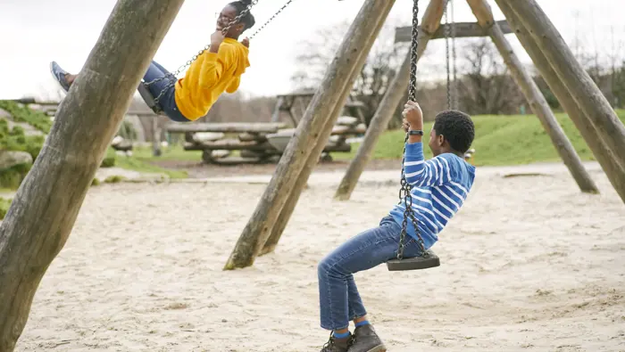 Young girl in yellow hoodie and boy in striped jumper swinging high at play area in Cuningar Loop Woodland Park, Glasgow
