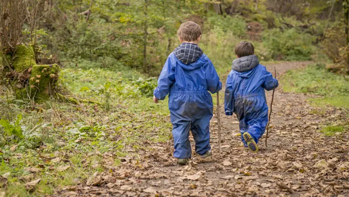 Two young boys in blue wet weather gear, each holding a stick, walk along forest path strewn with leaves, Rannoch Wood, Renfrewshire Woods, near Johnstone