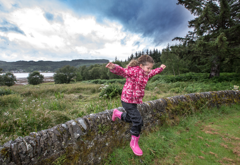 Young girl in pink jacket and wellington boots jumps from stone wall, with loch in background, Ardcastle Forest
