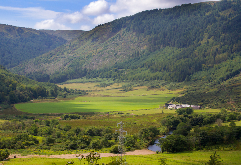 A semi wooded chain of hills with a valley of fields, houses, a small river, and power lines. 
