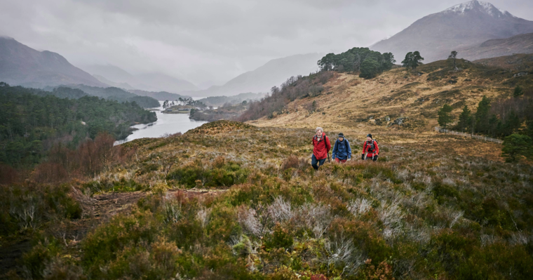 three people walking across scrubby terrain with mountains and a loch in the background