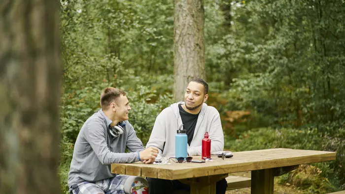Two men (one with prosthetic leg) sit together chatting at picnic table, on woodland trail, Devilla Forest, near Kincardine