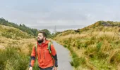 Man in orange jacket wearing haversack walking on coastal trail with loch in background, near Beinn Lora, Benderloch