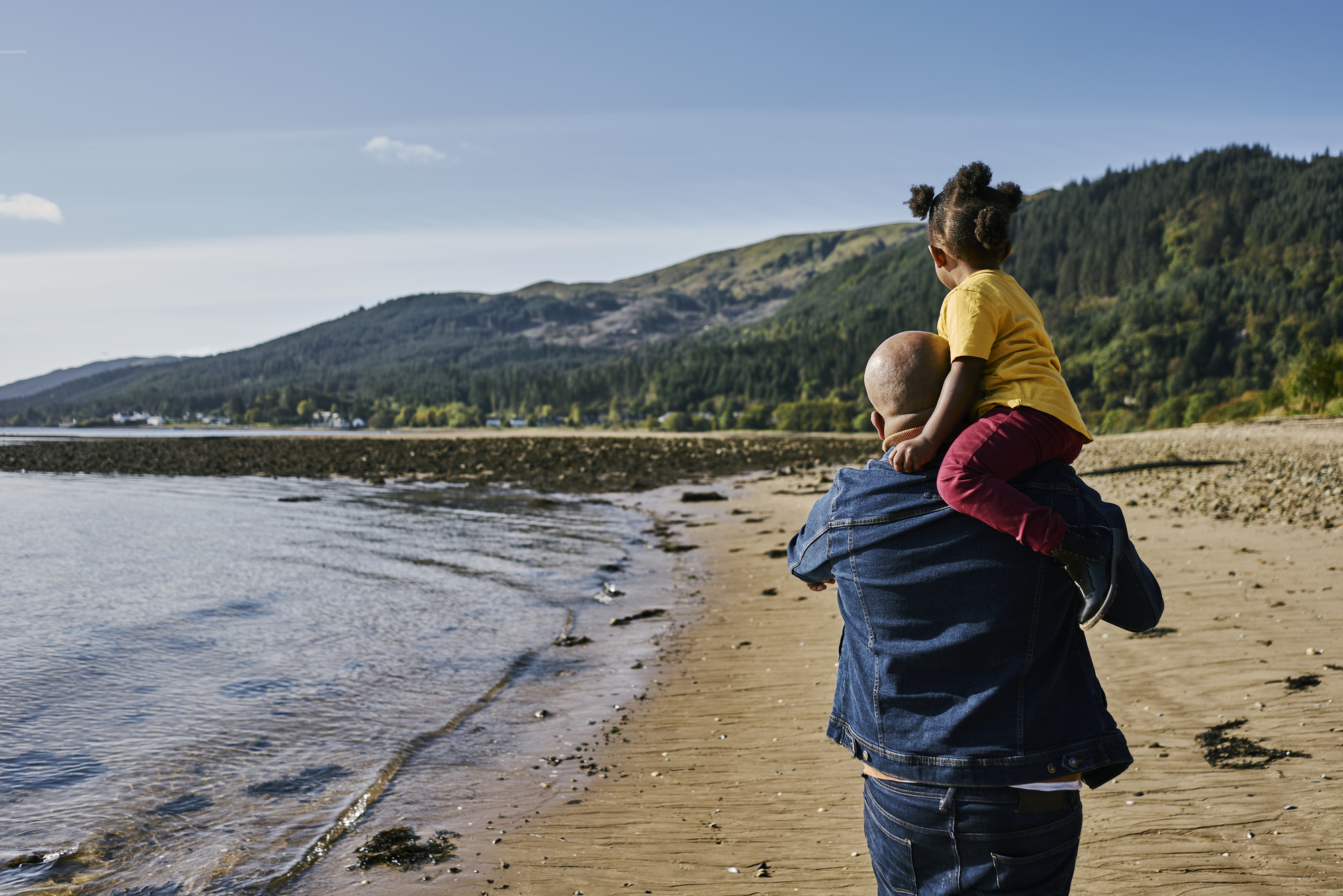 Man with young girl on shoulders walks along sandy Ardentinny beach on shores of Loch Long, with hills in background
