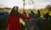 Rear view of woman in evening sun, wearing red jacket, touching boulders in Touchstone Maze, labyrinth of Scottish stones, Blackmuir Wood, near Strathpeffer