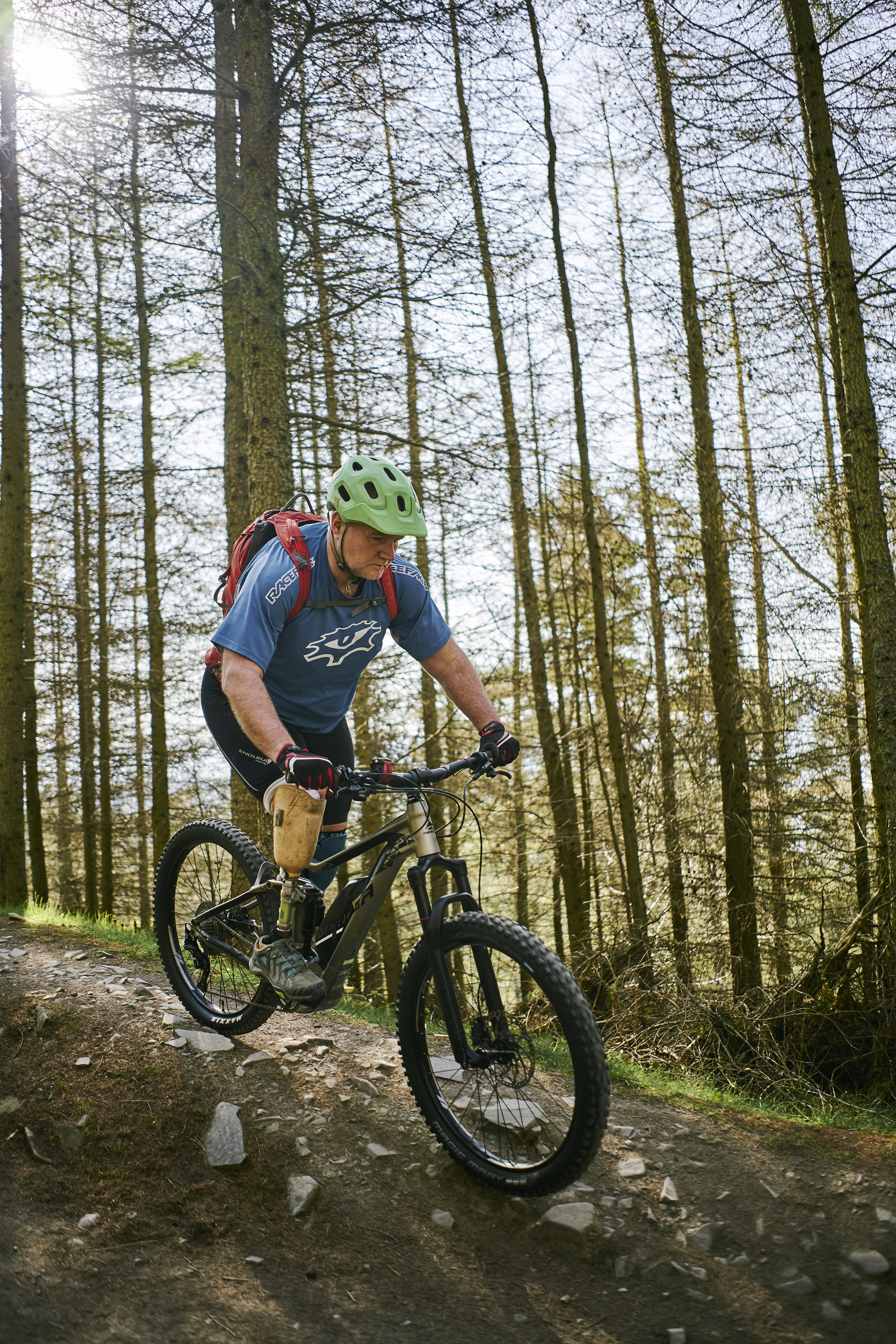 Male mountain biker (with prosthetic leg) wearing blue T shirt and green helmet rides bike along stony ridge flanked by a bank of tall trees, Glentress Forest, near Peebles