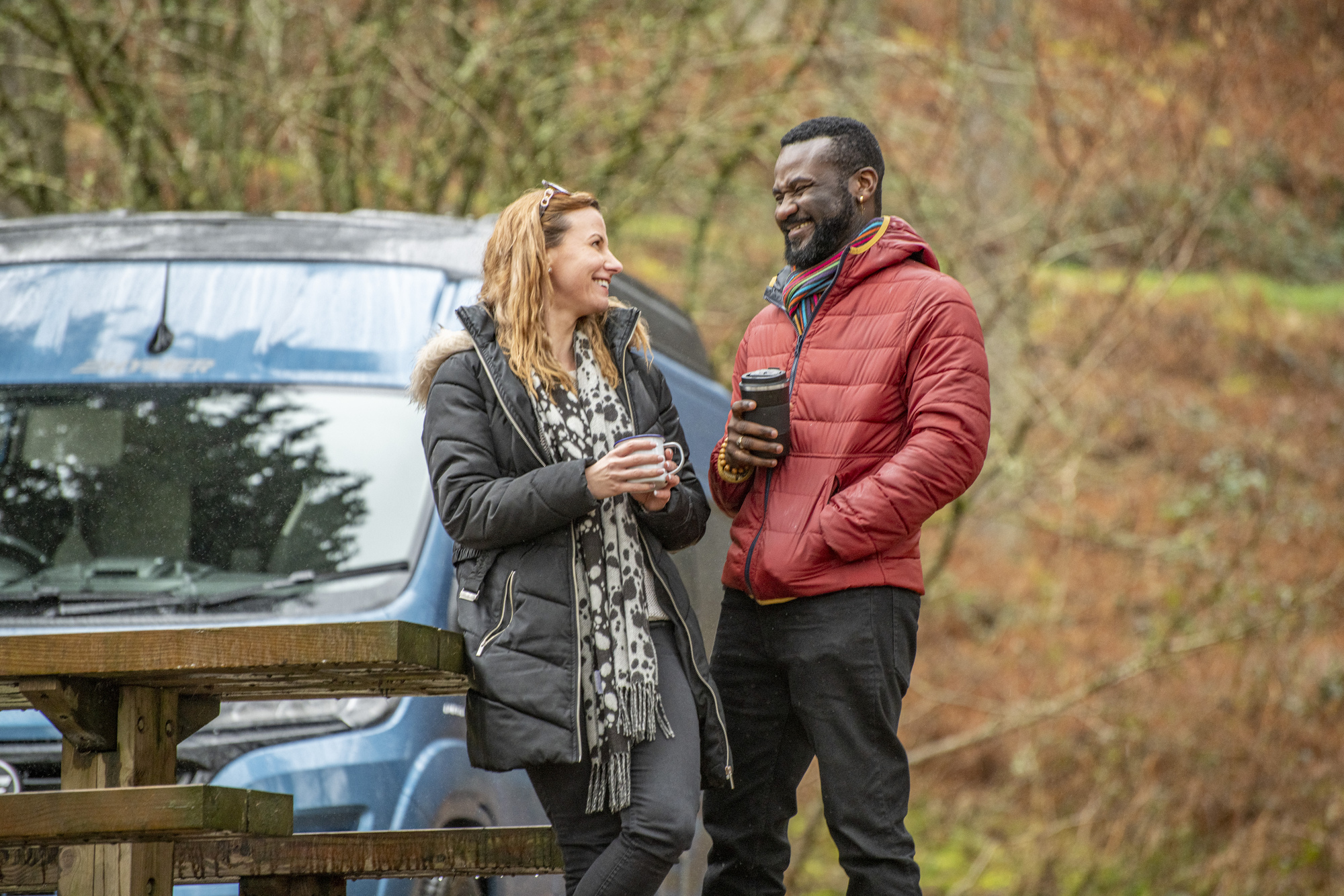 couple standing next to picnic table in front of campervan with reusable travel coffee mugs