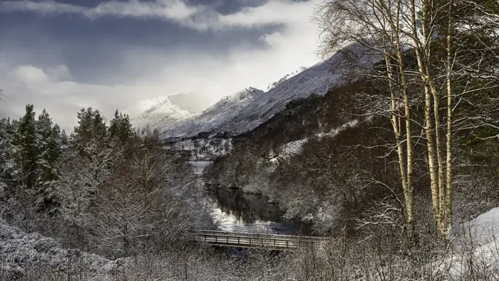 Glen with bridge in the foreground and mountains in dramatic light in the background.