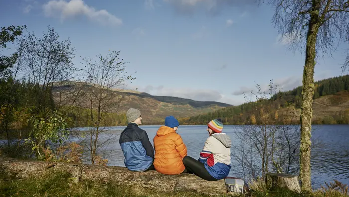 Three people sitting on a log bench by a loch.