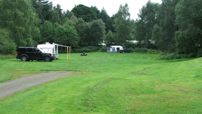 Green grassy field with gravel road through it and trees beyond, with a parked caravan