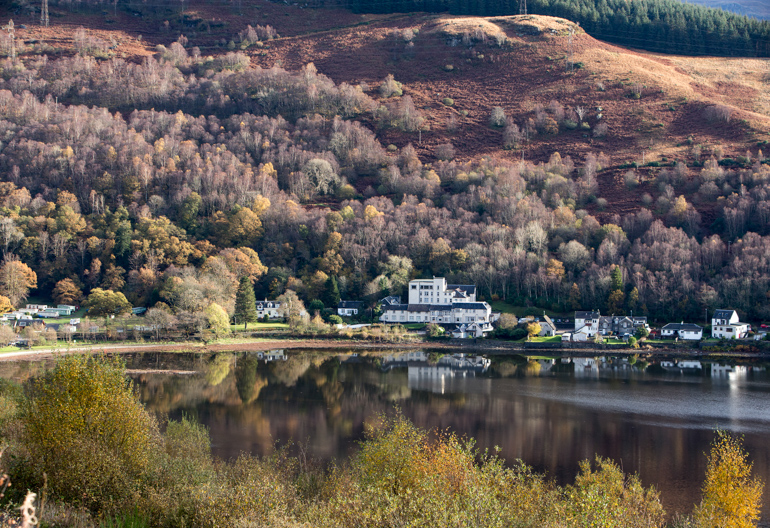 View across still water to a small village on opposite shore in front of a hillside of mixed trees and brush.