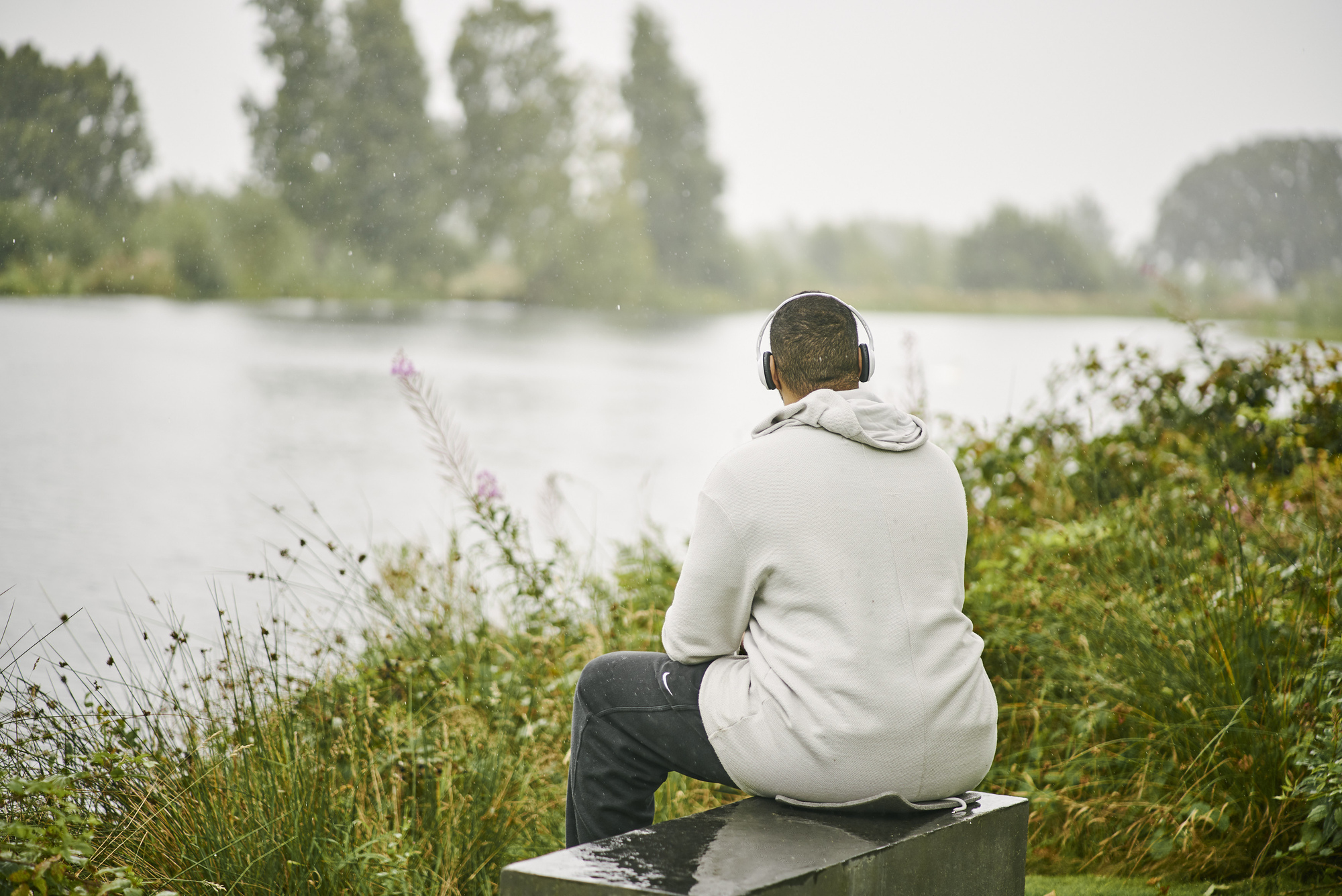 Man in white jacket wearing headphones sits on bench looking out at pond, Larbert Woods in grounds of Forth Valley Royal Hospital, Larbert