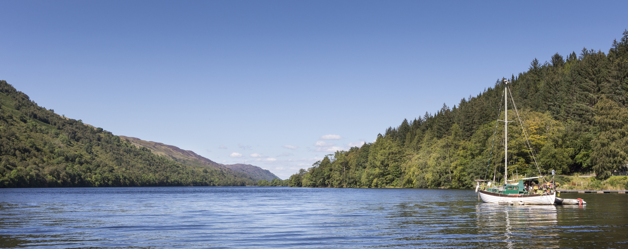 A sail boat tied up in calm water with a forested hillside in the background.