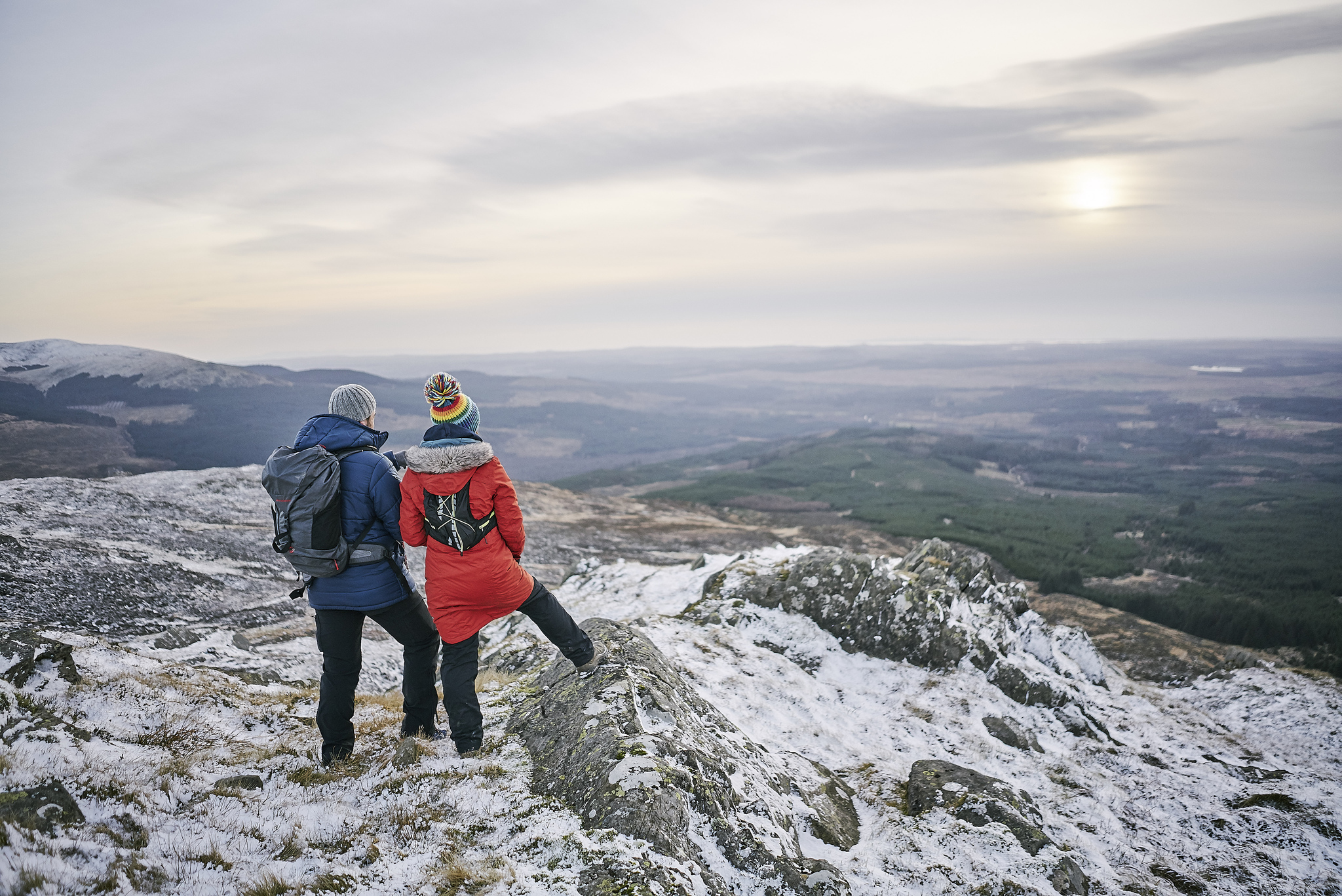 A couple standing on a snowy hillside with expansive views of forestry and farmland.