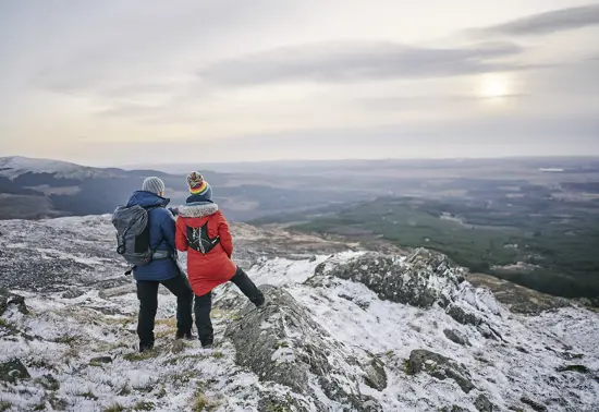 A couple standing on a snowy hillside with expansive views of forestry and farmland.