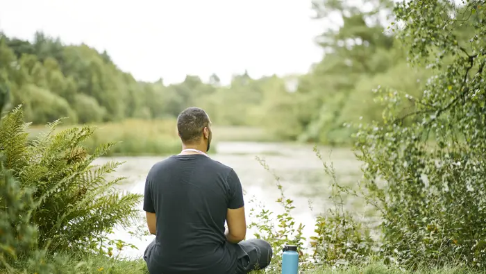 Man in black t shirt with blue flask beside him, sits on grass edge of Bordie Loch, Devilla Forest, near Kincardine