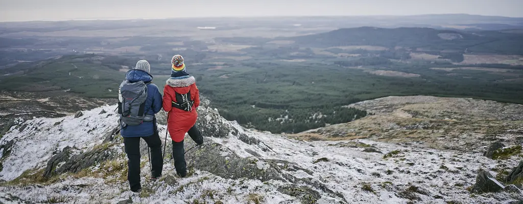 Two people standing on a hilltop looking out over hills and forests in winter.