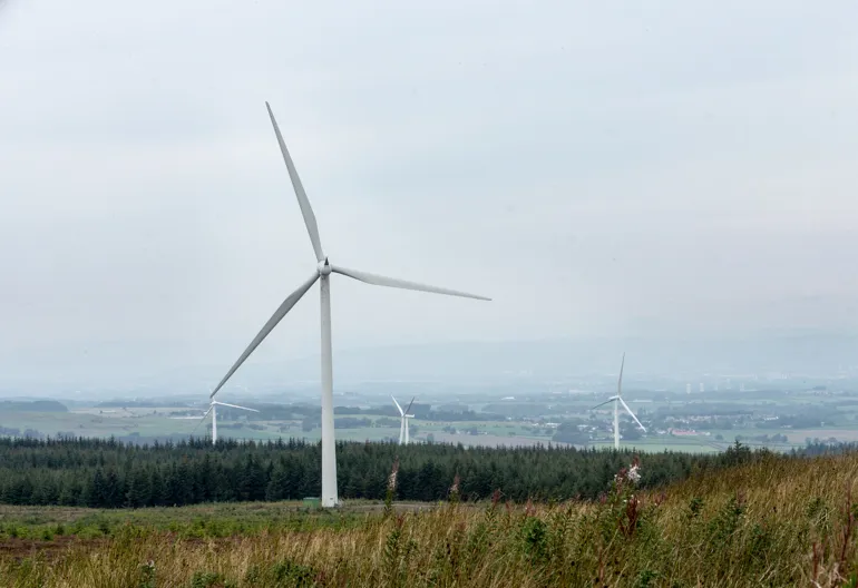 Large windmills in a windfarm with trees and fields.
