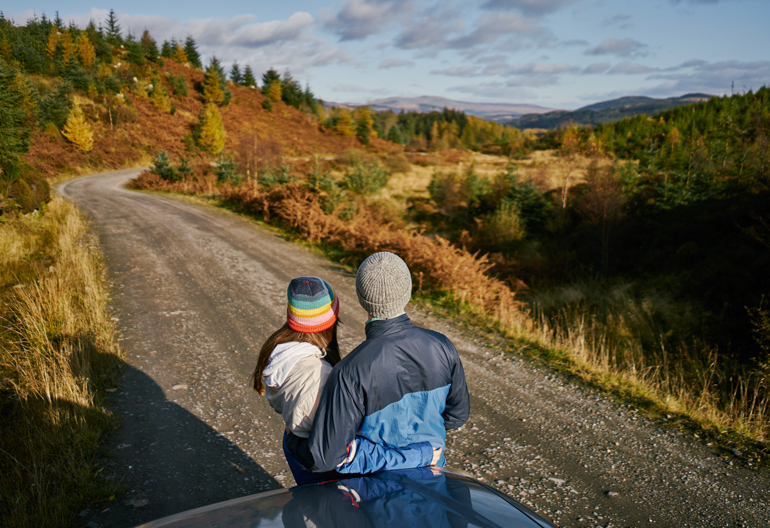 A woman in a rainbow hat and a man in a blue jacket lean against a vehicle on a forest road looking at the wooded valley below