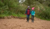 Woman and young girl walk hand in hand along the sand with woodland in background, at Boden Boo riverside beach, Renfrewshire Woods, near Erskine Bridge